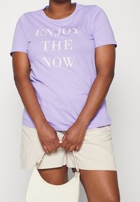 Lavendel bomulds t-shirt med "NYD NUET" trykt i hvid, parret med lyse beige shorts og en lille hvid håndtaske.
