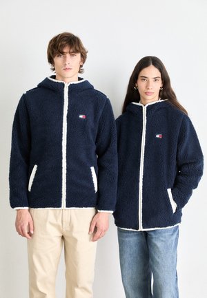 Sweatshirts en polaire bleu marine avec des accents blancs, tissu texturé et poches avant. Les modèles portent des jeans beige clair et bleus pour le contraste.