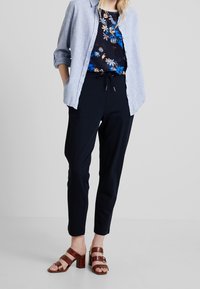 Hellblaues Button-up-Hemd über einer dunklen Blumenbluse, kombiniert mit schwarzen Hosen und braunen Absatzsandalen. Flache Oberfläche, neutraler Hintergrund.