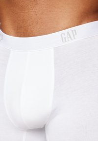 Boxer-chaussettes en coton blanc avec une ceinture élastique douce arborant le logo "GAP" en gris clair. Texture lisse et design ajusté.
