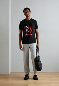 Black t-shirt with red and white graphic design, light grey trousers, black shoes, and a black bag. Simple background with wood flooring.