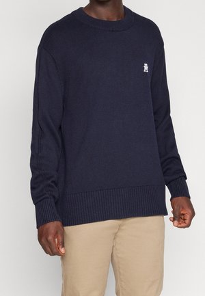 Mann trägt einen marineblauen Langarmpullover mit gerippten Bündchen und Saum, kombiniert mit beigen Hosen, stehend vor schlichtem Hintergrund.