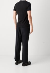 Černé polo tričko s bílými akcenty, spárované s širokými černými kalhotami. Materiál vypadá jemně; boty jsou hladké a černé. Zobrazeno z pohledu zezadu.