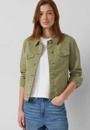 Jonge vrouw met rood haar, gekleed in een olijfgroen jack met knopen over een wit shirt, met blauwe jeans en één hand in de zak.