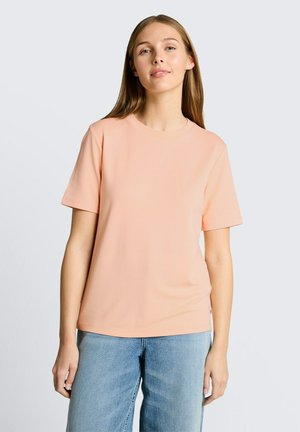 Giovane donna che indossa una maglietta pesca a maniche corte e jeans azzurri, in piedi davanti a uno sfondo grigio chiaro uniforme.