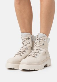 Bottines beige en suède, avec quincaillerie en argent, un col rembourré et une semelle plate-forme épaisse. Les lacets sont bien visibles.