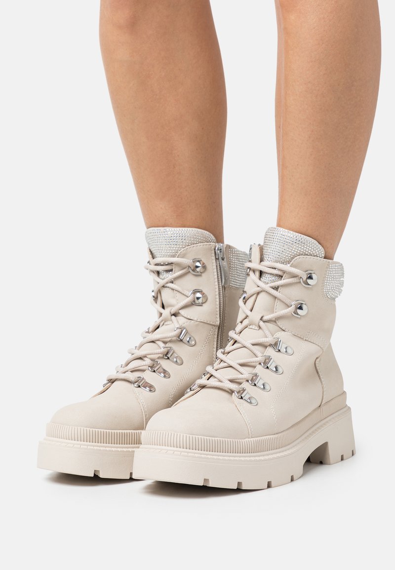 Bottines beige en suède, avec quincaillerie en argent, un col rembourré et une semelle plate-forme épaisse. Les lacets sont bien visibles.