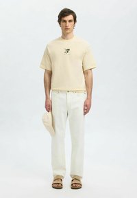 Kremowa bawełniana koszulka z krótkim rękawem i małym nadrukiem, w połączeniu z jasnobeżowymi dżinsami, dopasowaną czapką i brązowymi sandałami.