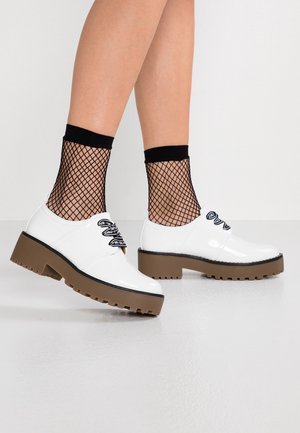 Witte schoenen van lakleer met zwarte veters, plateauzolen en een bruine, geribbelde zool. Draagt zwarte visnet enkelsokken.