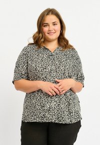 Camicia con stampa leopardata nera e beige, colletto, maniche corte e vestibilità ampia. Il tessuto sembra leggero e ha una texture liscia.