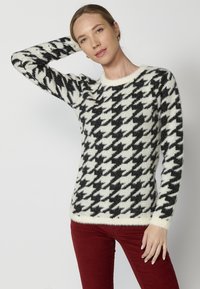 Pulover cu model houndstooth alb-negru, realizat dintr-un material moale și pufoș, având mâneci lungi și un guler rotund.