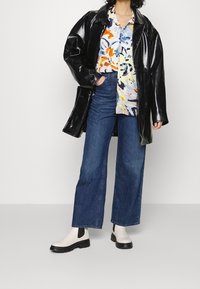 Osoba w kolorowej koszuli z abstrakcyjnym wzorem, czarnym, błyszczącym skórzanym płaszczu, szerokich niebieskich dżinsach oraz białych botkach za kostkę na jednolitym tle.