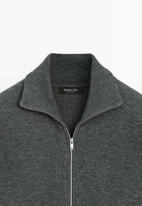 Maglione grigio lavorato a maglia con colletto zip. Presenta una texture liscia e dettagli a coste sul colletto e sui polsini. Etichetta del marchio visibile all'interno.