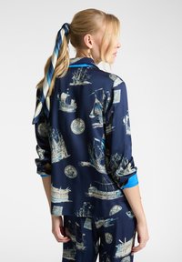 Camicia di seta blu navy con stampe di navi, caratterizzata da un colletto classico e maniche arrotolate, abbinata a pantaloni coordinati.