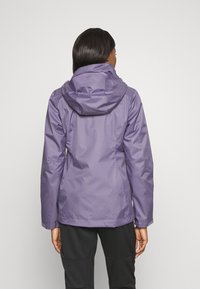 Femme de dos portant une veste imperméable à capuche lavande et un pantalon noir texturé, sur un fond uni.