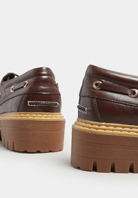 Ruske nahkasaappaat paksulla ruskealla pohjalla, joissa on kultaiset silmukat, koristeellinen ommel ja teksturoitu pinta kannassa.