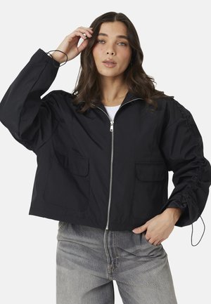 Vrouw met lang bruin haar die een zwart jasje met rits draagt met grote voorzakken en geplooide mouwen, gecombineerd met lichtgrijze spijkerbroek.