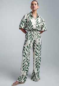 Krem jumpsuit s uzorcima zelenog lišća, kratkim rukavima, dugmetom na kopčanju i elastičnim strukom. Široke hlače i lagana teksturirana tkanina.