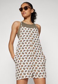 Witte romper met een meerkleurig bloemen- en cactuspatroon, mouwloos ontwerp, zijzakken en verstelbare banden. Bruine zonnebril accentueert.