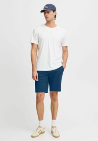 Hvitt bomull t-skjorte, blå shorts, beige joggesko med hvite detaljer, og en marineblå caps med tekst. Modell står med hendene i lommene.