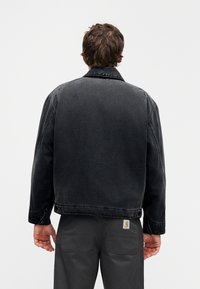 Μαύρο τζιν μπουφάν με γιακά, εμφανείς ραφές και κλείσιμο με κουμπιά. Απλός σχεδιασμός, φορεμένο πάνω από γκρι παντελόνια.