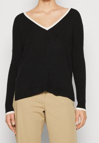 Maglione nero a maniche lunghe con scollo a V e bordi bianchi a contrasto sul colletto e sui polsini. Tessuto morbido, vestibilità rilassata e orlo leggermente curvo.