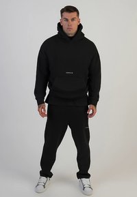 Musta huppari- ja collegehousupuku, valmistettu pehmeästä kankaasta. Hupparissa on kengurutasku ja etupuolella logo; collegehousuissa on hillitty koristeellinen yksityiskohta.