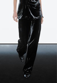 Person walking in shiny black velvet pants and matching top, with visible arms and shoes on a gradient floor.