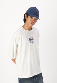Hvid oversized t-shirt med et centralt grafikprint og korte ærmer. Sammen med en mørkeblå baseball cap. Bomuldsstof, afslappet design.