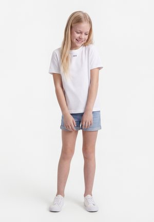 Κορίτσι με μακριά ξανθά μαλλιά, που φοράει λευκό μπλουζάκι με το κείμενο "OFF", τζιν σορτς και λευκά αθλητικά, χαμογελάει και κοιτάζει προς τα κάτω.