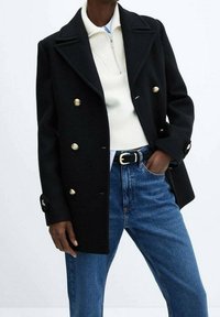 Abrigo negro cruzado con botones dorados, cuello con muesca y bolsillos laterales, superpuesto a un suéter blanco con cremallera y jeans azules.