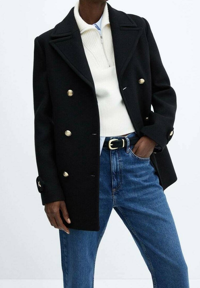 Abrigo negro cruzado con botones dorados, cuello con muesca y bolsillos laterales, superpuesto a un suéter blanco con cremallera y jeans azules.