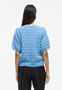 Jersey azul de punto con mangas abullonadas, que presenta una superficie texturizada de puntos blancos en relieve y un dobladillo acanalado. Sin cierres visibles.