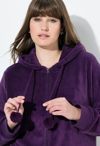 Sweat-shirt zippé violette en polaire avec capuche, doté de cordons de serrage à pompons et de poignets côtelés. Texture douce et coupe décontractée.