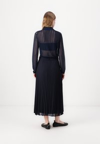Fustă plisată de culoare bleumarin asociată cu o bluză transparentă cu dungi. Bluza are mâneci lungi și guler, evidențiind o textură stratificată. Pantofi plat negri.
