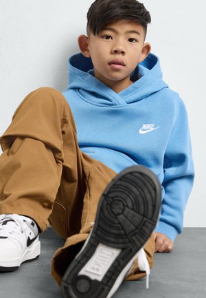 Sudadera de Nike azul claro con bolsillo canguro y logo blanco, combinada con pantalones marrones y zapatillas blanco y negro. Enfocada en ropa casual.