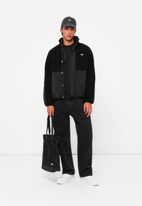 Zwarte fleecetrui met doorstikte panelen, zwarte jeans, witte sneakers en een zwarte pet, terwijl hij een zwarte tote bag met logodetails vasthoudt.