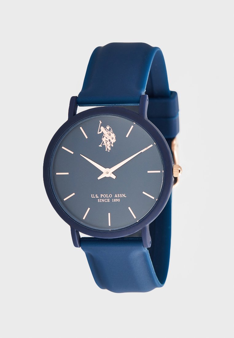Montre avec bracelet en silicone bleu marine, un cadran rond bleu marine, des index dorés et un logo. Design minimaliste avec des aiguilles simples pour les heures et les minutes.