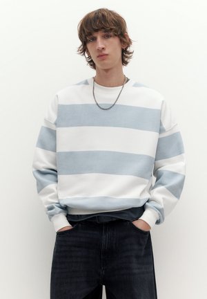 Mlad moški z razmršenimi rjavimi lasmi, oblečen v belo in svetlomodro široko črtasto jopico, črne kavbojke in srebrno verižico na vratu.