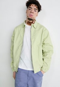 Jachetă verde deschis cu fermoar, guler și manșete elastice, având un logo subtil. Purtați peste un tricou alb și asortați cu pantaloni mov deschis.