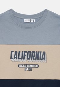 Majica v barvnih blokih z modrimi, bež in mornarskimi секцијami, з značko "CALIFORNIA Original Urban Brand EST. 1986" na prsih.