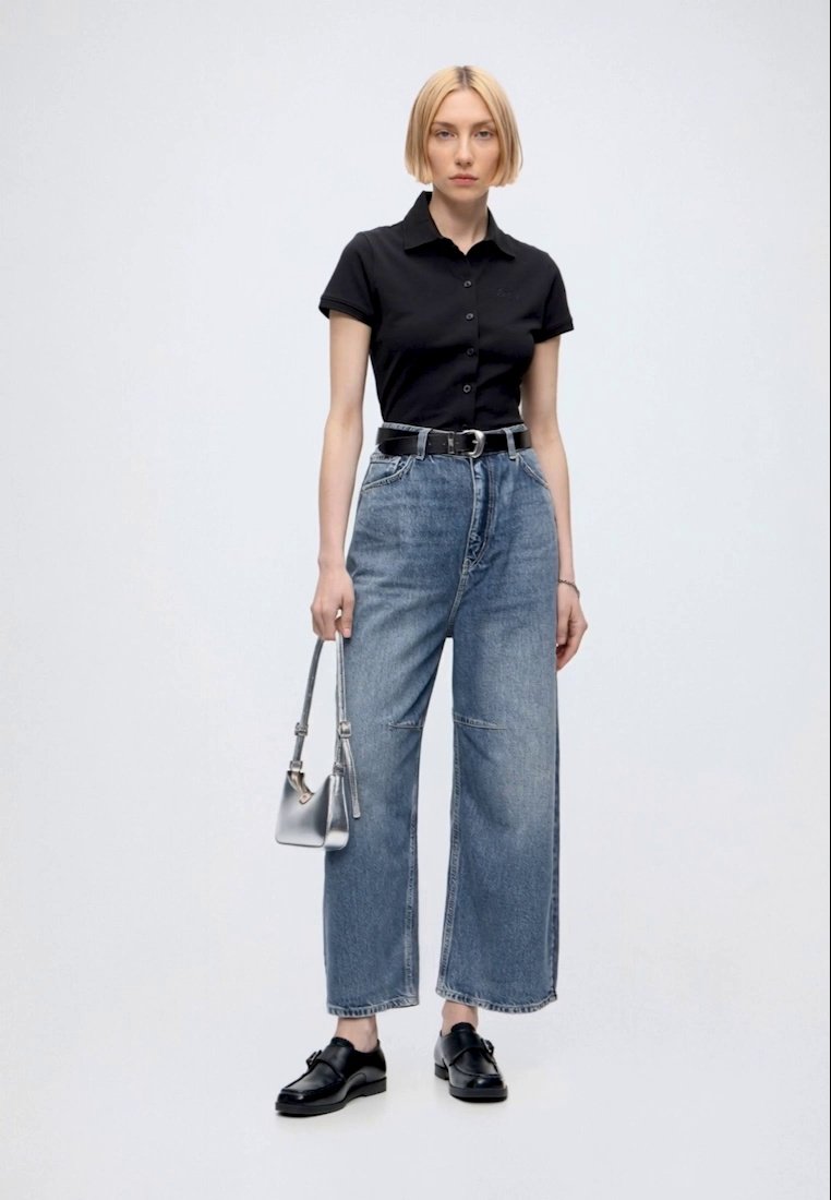 Femme portant une chemise noire à manches courtes avec des boutons, un jean bleu à jambes larges, une ceinture noire, des chaussures noires et tenant un petit sac à main argenté.