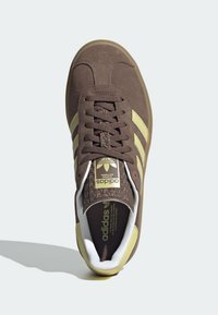 Brown suede Adidas sneaker with beige stripes, brown laces, and patterned tongue, viewed from above on a white background.