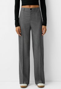 Pantalons larges gris à rayures avec fermeture à bouton et passants de ceinture. Le matériau a une texture lisse. Associés à des chaussures de couleur claire.