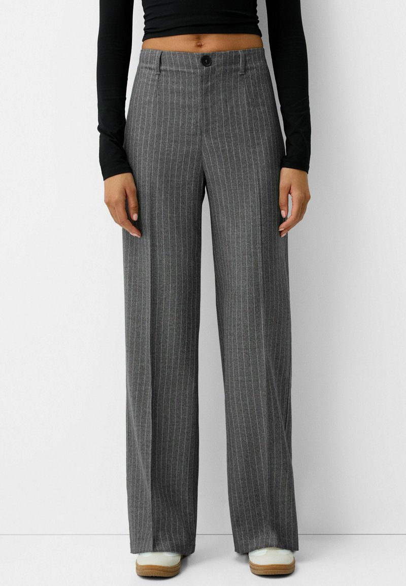 Pantalons larges gris à rayures avec fermeture à bouton et passants de ceinture. Le matériau a une texture lisse. Associés à des chaussures de couleur claire.