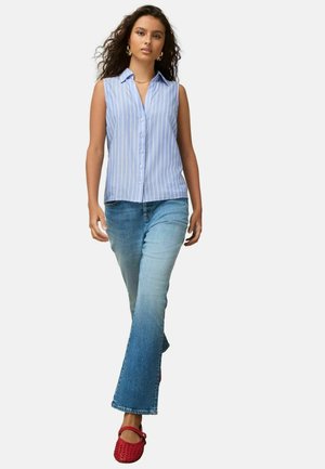 Kobieta idąca, ubrana w niebiesko-białą pasiastą koszulę bez rękawów na guziki, wyblakłe niebieskie dżinsy, czerwone plecione buty oraz złote kolczyki koła.