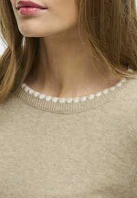 Suéter beige con cuello redondo, adornado con un ribete de pompón blanco. La tela parece suave, con una textura lisa y un sutil patrón jaspeado.
