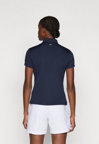 Camisa polo azul-marinho com mangas curtas e colarinho, feita de tecido liso, com um pequeno logótipo em azul claro nas costas. Calções brancos.