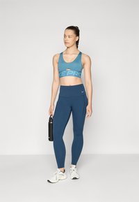 Športni nedrček v teal barvi z ovalnim izrezom in blagovno znamko "NIKE PRO". Ujemajoče se visokorasle legice v temno modri barvi. Črna steklenica za vodo v roki.