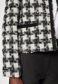 Gros plan sur la manche et la poche d'une veste en tweed texturée noir et blanc, portée sur un pantalon noir, mettant en valeur les détails du tissu et le motif.